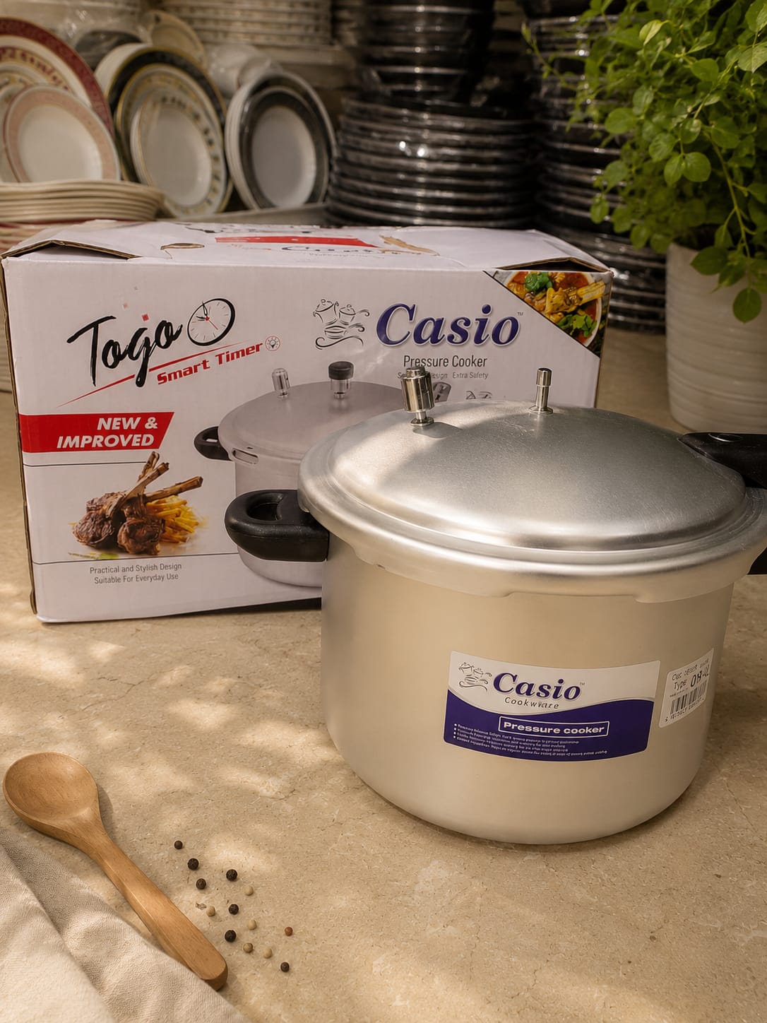 Casio aluminium pressure cooker photographed in front of its printed box with stacked crockery behind